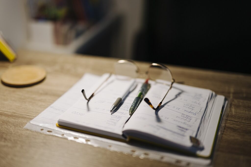 A desk with an open notebook, pens and a pair of glasses